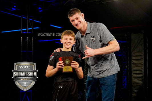 Zachary Helka holding his winner's plaque after a submission victory at WXC Submission Series, standing beside a coach inside the cage.